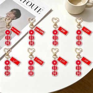 Pode incluir: Uma coleção de chaveiros vermelhos e brancos com fechos dourados em forma de coração. Cada chaveiro apresenta uma série de três círculos vermelhos e uma etiqueta retangular com um nome ou texto, incluindo "BTS WORLD TOUR", "JUNG KOOK" e "JIMIN".