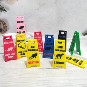 Könnte beinhalten: Eine Sammlung farbenfroher Kunststoff-Warnschilder mit humorvollen Warnungen zu Katzen und Hunden. Die Schilder sind rot, gelb, rosa, blau, schwarz und grün, mit Text und grafischen Illustrationen. Auf den Schildern steht "Katzenkotze", "Hunde-Durchfall".