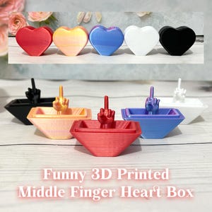Può includere: Una collezione di scatole a forma di cuore e oggetti a forma di cuore stampati in 3D in vari colori, tra cui rosso, giallo, blu, bianco e nero. Ogni scatola presenta un gesto del dito medio. Il testo "Funny 3D Printed Middle Finger Heart Box" è visualizzato.