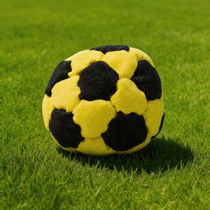 Op de afbeelding: Een zachte, pluche voetbal met een klassiek ontwerp. De bal heeft een patroon van zwarte en gele vijfhoeken, tegen een achtergrond van groen gras. Dit speelgoed is geschikt voor binnen- of buitenspelen.