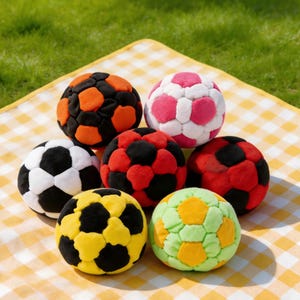 Op de afbeelding: Een verzameling van zeven kleurrijke voetballen met verschillende kleurencombinaties, waaronder zwart en oranje, roze en wit, zwart en wit, rood en zwart, geel en zwart en groen en geel. De ballen zijn gerangschikt op een geel-wit geblokt picknickkleed.