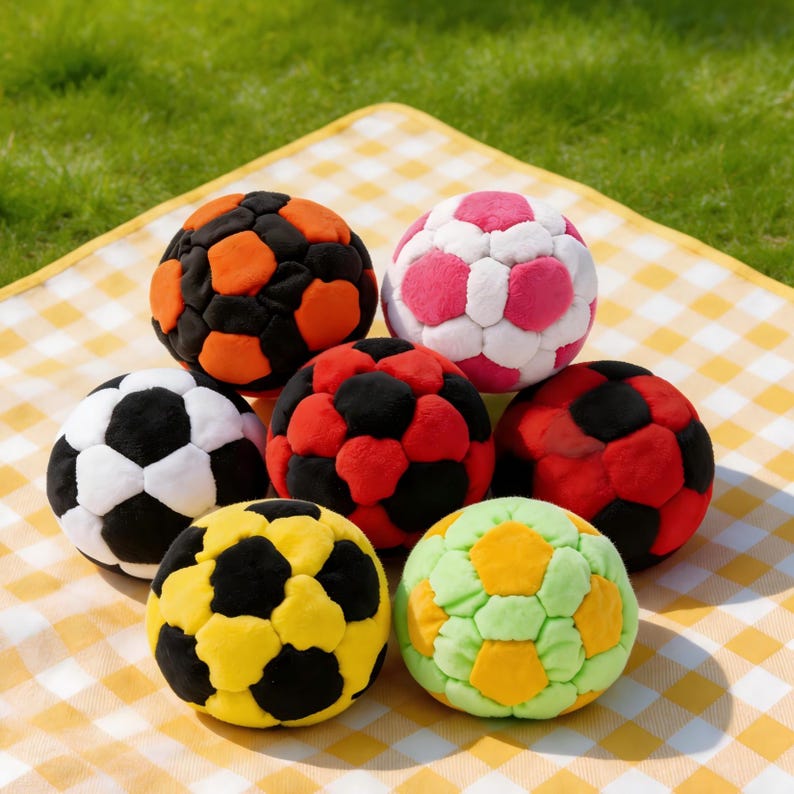 Op de afbeelding: Een verzameling van zeven kleurrijke voetballen met verschillende kleurencombinaties, waaronder zwart en oranje, roze en wit, zwart en wit, rood en zwart, geel en zwart en groen en geel. De ballen zijn gerangschikt op een geel-wit geblokt picknickkleed.