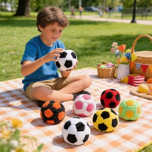 Op de afbeelding: Een kind houdt een zwart-witte voetbal vast. Verschillende kleurrijke voetballen in oranje, roze, rood, geel en groen zijn gerangschikt op een picknickkleed. De sc&egrave;ne speelt zich buiten af op een zonnige dag.