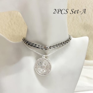 May include: A two-piece silver necklace set displayed on a white mannequin. The set includes a chunky chain necklace and a delicate chain with a round pendant. The pendant is intricately detailed with a floral design and small, sparkling accents. The text "2PCS Set-A" is visible.
