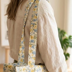 May include: A floral tote bag with a cream background and a pattern of blue and yellow flowers. The bag has matching straps and is being carried over a person's shoulder. The person is wearing a beige linen shirt.