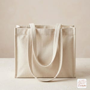 May include: A rectangular, off-white canvas tote bag with two long straps. The bag is made of a sturdy, textured fabric and has a simple, minimalist design. The straps are long enough to be worn over the shoulder. The bag is empty.