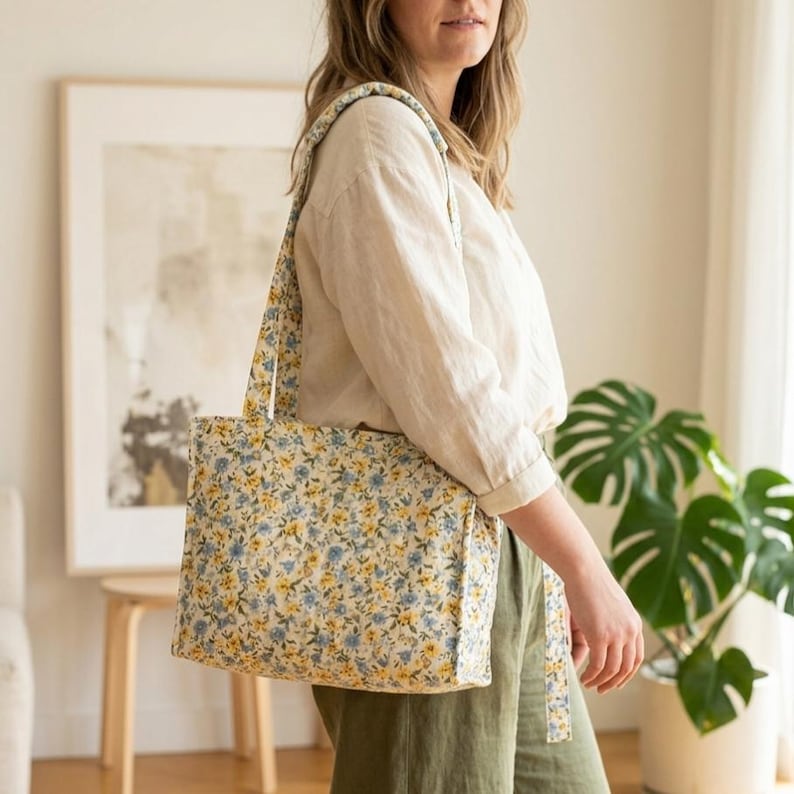 May include: A floral print tote bag with a rectangular shape and long straps. The bag features a pattern of small blue and yellow flowers on a cream background. The straps match the bag's floral design. The bag is carried over the shoulder.
