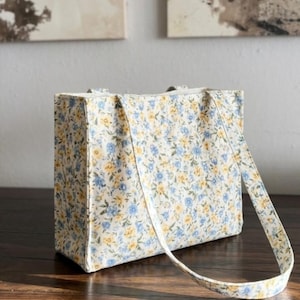 May include: A rectangular tote bag with a floral pattern in blue and yellow on a cream background. The bag has a matching strap and is sitting on a wooden surface.