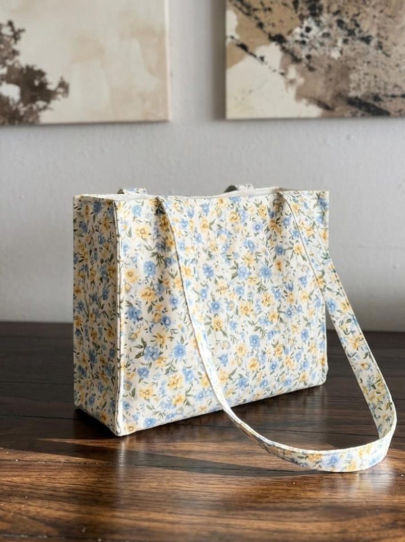 May include: A rectangular tote bag with a floral pattern in blue and yellow on a cream background. The bag has a matching strap and is sitting on a wooden surface.