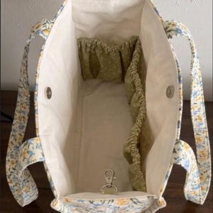 May include: Open tote bag with a floral pattern in blue, yellow, and white. The interior is cream-colored with olive green pockets. A silver-colored clasp is visible at the bottom. The handles are floral patterned.