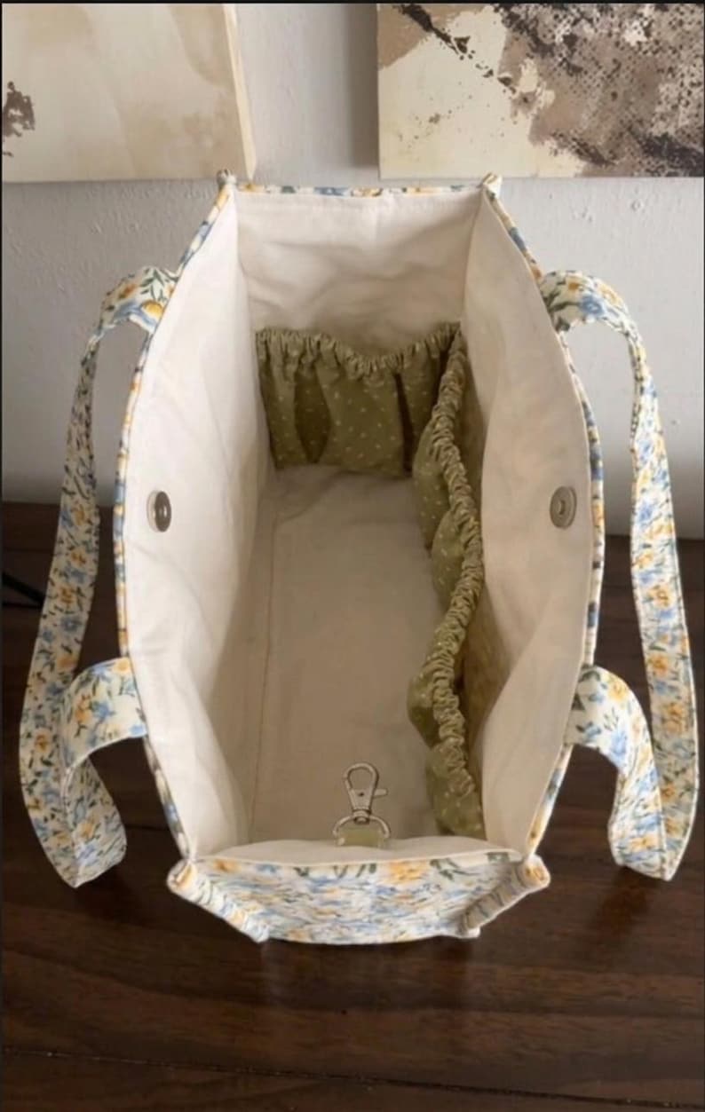 May include: Open tote bag with a floral pattern in blue, yellow, and white. The interior is cream-colored with olive green pockets. A silver-colored clasp is visible at the bottom. The handles are floral patterned.