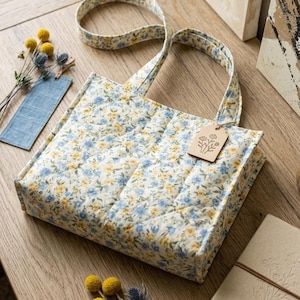 May include: A quilted tote bag with a floral pattern in blue and yellow on a cream background. The bag has matching handles and a wooden tag. The bag is sitting on a wooden surface with decorative items.