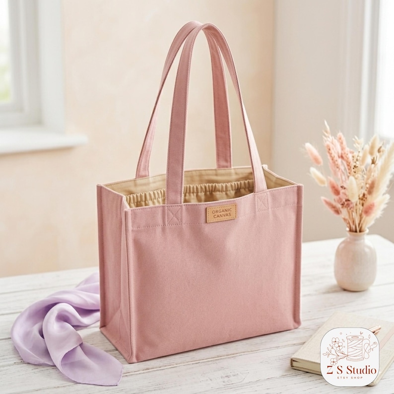 May include: A pink canvas tote bag with matching handles. The bag has a rectangular shape and a light tan interior. A small, rectangular tag with the words "ORGANIC CANVAS" is sewn on the front. A purple scarf and a small vase of dried flowers are nearby.