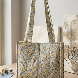 May include: A quilted tote bag with a floral pattern in yellow and blue on a cream background. The bag has two handles and a rectangular shape. The bag is sitting on a wooden surface.