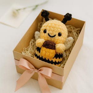 Peut inclure: Un jouet en peluche fait main en forme d'abeille, jaune et noir, avec un visage souriant, des antennes noires et un pot de miel marron, présenté dans une boîte cadeau marron avec un ruban rose.