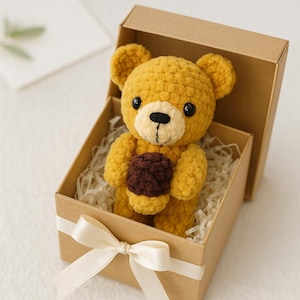 Könnte beinhalten: Ein handgefertigter gelber Häkelteddybär, der eine braune Eichel hält, präsentiert in einer braunen Geschenkbox mit weißem Band. Der Bär hat schwarze Augen und eine cremefarbene Schnauze, eingebettet in geschreddertes Papier.