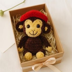 May include: A handmade crochet monkey doll with a red beret, presented in a gift box with a ribbon. The monkey has brown fur, a tan face, and yellow accents on its hands and feet. The box is filled with shredded paper.