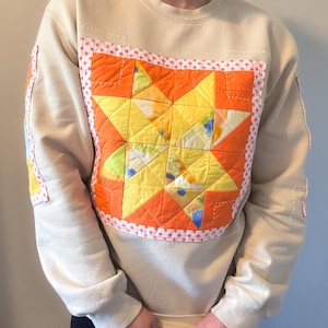 May include: A beige crewneck sweatshirt featuring a large, central quilted star patch in vibrant orange and yellow tones. The star is framed by a white fabric border with a small red floral pattern, and similar quilted patches are visible on the sleeves.