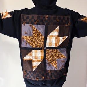 Peut inclure: Un sweat-shirt à capuche noir avec un grand empiècement matelassé carré dans le dos. L'empiècement présente un motif d'étoile à quatre branches dans des tons marron, beige et gris, avec des bordures à carreaux et de petits liens effilochés.