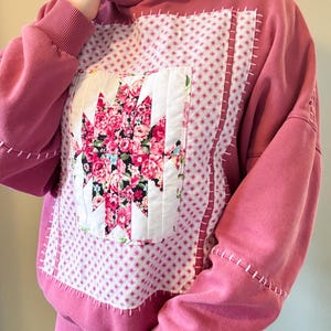 May include: A dusty rose hooded sweatshirt featuring a large rectangular patch on the front. The patch is white with a small pink star pattern and a central quilted floral block in shades of pink, white, and black. Visible pink hand-stitched detailing.