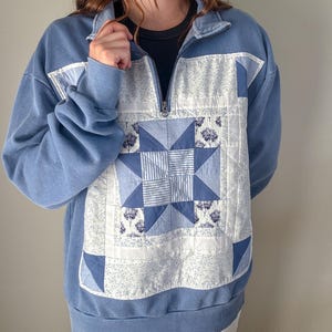 Pode incluir: Uma camisola azul com fecho de um quarto, apresentando um grande painel frontal acolchoado e quadrado. O painel exibe um padrão de estrela central em tons de azul e branco, rodeado por blocos de tecido floral e às riscas, com punhos canelados.