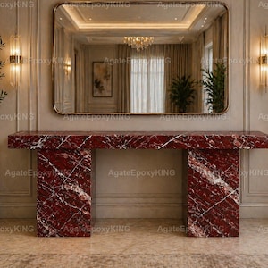 Puede incluir: Una consola rectangular de mármol rojo con vetas blancas. La mesa tiene una parte superior gruesa y dos pilares de soporte. Un gran espejo con marco dorado cuelga sobre la mesa, reflejando una lámpara de araña y detalles interiores. La mesa es una pieza destacada.