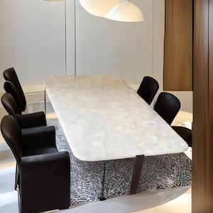 Puede incluir: Un juego de comedor moderno con una mesa rectangular con efecto mármol y patas de metal oscuro. La mesa está rodeada de sillas de cuero negro. La base de la mesa está cubierta con una tela estampada. Tres lámparas colgantes blancas iluminan la escena.