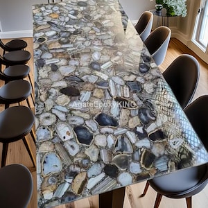 Könnte beinhalten: Ein rechteckiger Esstisch mit einer polierten Oberfläche aus eingelegten Steinen in Grau-, Blau- und Beigetönen. Der Tisch ist von schwarzen Stühlen umgeben. Der Text "AgateEpoxyKING" ist auf der Tischoberfläche sichtbar.
