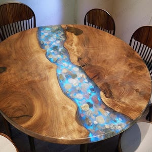 May include: Round wooden table with a river-like design. The center features a blue epoxy resin filled with stones, creating a unique, illuminated effect. The table is surrounded by wooden chairs.
