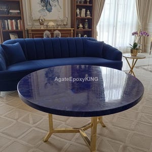 Può includere: Un tavolino rotondo blu navy con una base in metallo color oro. Il tavolo ha una superficie lucida e marmorizzata. Il testo "AgateEpoxyKING" è visibile sul tavolo. Un divano blu navy è sullo sfondo.