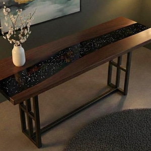 Puede incluir: Una consola de madera oscura con un diseño de río de resina epoxi negra incrustado en el centro. La mesa tiene forma rectangular con patas de metal. Un jarrón blanco con flores blancas está sobre la mesa.