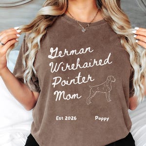 May include: A brown t-shirt with the text "German Wirehaired Pointer Mom" in white script. Below the text is a line drawing of a dog. The shirt also includes the text "Est 2026" and the name "Poppy".