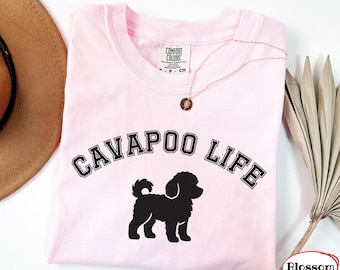 Peut inclure: Un t-shirt rose clair à manches courtes avec le texte noir CAVAPOO LIFE en police universitaire arqué au-dessus de la silhouette noire d'un chien aux poils bouclés. L'étiquette intérieure du col indique COMFORT COLORS et la taille S-P-CH.