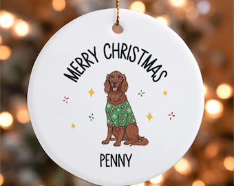 Op de afbeelding: Een witte, ronde kerstversiering met de tekst "MERRY CHRISTMAS" in het zwart. De versiering toont een bruine hond met een bril en een groene trui met zuurstokontwerpen. De naam van de hond, "PENNY", staat eronder afgedrukt.