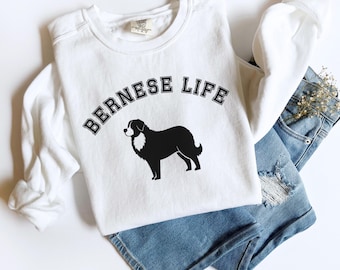 Puede incluir: Sudadera blanca de cuello redondo con el texto "BERNESE LIFE" sobre una silueta en blanco y negro de un perro de montaña bernés. La sudadera se combina con pantalones cortos vaqueros azules.