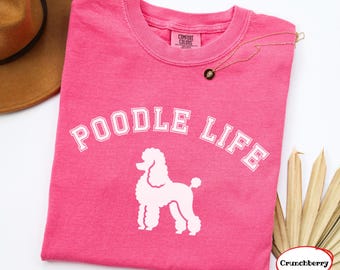 May include: A bright pink short-sleeved t-shirt featuring the white text POODLE LIFE in an arched collegiate font above a white graphic of a standing poodle. The shirt has a crew neckline and a Comfort Colors brand label on the inner collar.