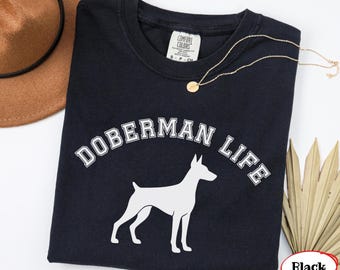 Può includere: Una t-shirt nera a maniche corte con una grafica bianca della sagoma di un cane Doberman e il testo DOBERMAN LIFE in un carattere universitario. La maglietta presenta un'etichetta del marchio Comfort Colors sul colletto interno.