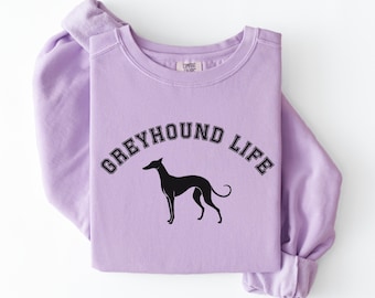 May include: A light purple sweatshirt with the words "GREYHOUND LIFE" arched above a black silhouette of a greyhound dog. The sweatshirt is folded, showing the sleeves and collar.