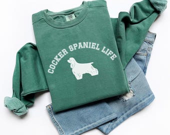 May include: A green sweatshirt with the text "COCKER SPANIEL LIFE" and a white Cocker Spaniel graphic. The sweatshirt is paired with light blue denim jeans with distressed details. The items are arranged on a white surface.