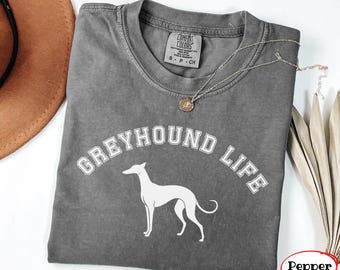 May include: A charcoal gray short-sleeve t-shirt featuring a white graphic of a greyhound dog and the text "GREYHOUND LIFE" in a collegiate font. The shirt has a Comfort Colors brand label at the inner neckline and a thin gold-toned necklace.