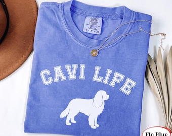 Puede incluir: Una camiseta de manga corta en color azul bígaro con un estampado gráfico blanco de la silueta de un perro y el texto CAVI LIFE en una fuente universitaria. La camiseta presenta una etiqueta de la marca Comfort Colors en el cuello.