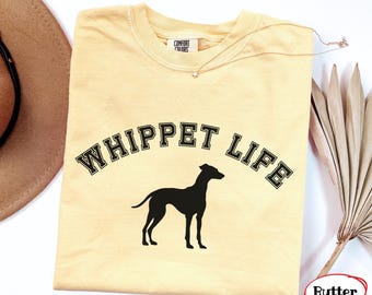 May include: A pale yellow short-sleeve t-shirt featuring the black text WHIPPET LIFE in a collegiate font arched over a black silhouette of a whippet dog. The shirt has a Comfort Colors brand label at the inner neckline and a crew neck collar.
