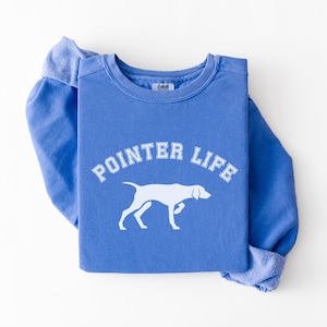 May include: A blue sweatshirt featuring the text "POINTER LIFE" in white and a white dog silhouette. The sweatshirt is folded, with the sleeves showing. The design is a tribute to dog lovers.