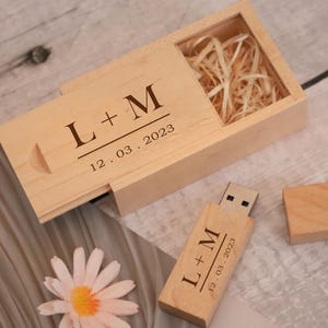 Può includere: Una chiavetta USB e una scatola di legno con le iniziali "L + M" e la data "12.03.2023" incise. La scatola aperta mostra della paglia. Una margherita è posizionata accanto.