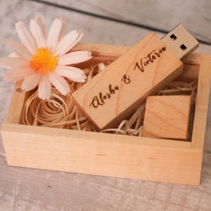 Puede incluir: Una memoria USB de madera grabada con "Aleisha & Victoria" descansa en una pequeña caja de madera llena de paja. Una margarita rosa claro y amarilla está en la caja. La caja es de color marrón claro.