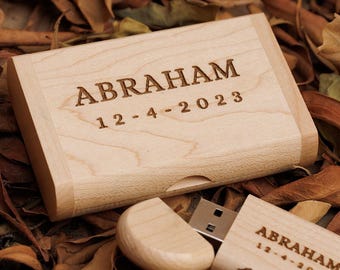 May include: A light-colored wooden USB flash drive and its matching case. The case and drive are engraved with the name "ABRAHAM" and the date "12-4-2023". The items are set against a backdrop of dried leaves.