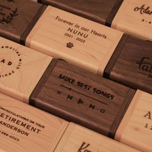 May include: Wooden boxes with engraved text and designs. The boxes are in shades of brown and beige. Some boxes feature phrases like "Forever in our Hearts" and "Congratulations on your Retirement". Other boxes have names and dates.