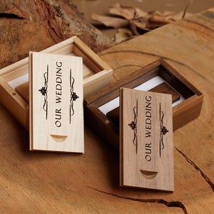 Peut inclure: Deux boîtes en bois pour clés USB, une claire et une foncée, avec l'inscription "OUR WEDDING" gravée sur le devant. Chaque boîte est ouverte, révélant la clé USB à l'intérieur. Les boîtes sont sur une surface en bois.