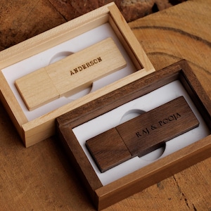 Op de afbeelding: Twee houten USB-sticks in bijpassende dozen. Eén is lichtgekleurd met "ANDERSON" gegraveerd, de andere is donkergeleurd met "RAJ & POOJA" gegraveerd. Beide zitten in wit gevoerde dozen.