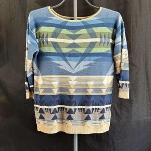 May include: A long-sleeve knit sweater featuring a horizontal geometric pattern in shades of blue, green, and beige. The garment has a round neckline, ribbed cuffs, and a ribbed hem, showcasing a repeating triangular and linear design throughout.